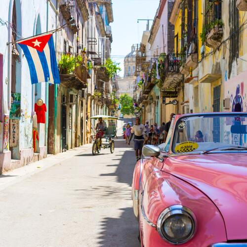 Een roze oldtimer staat in een smalle straat met oude huizen en een Cubaanse vlag in Havana, Cuba
