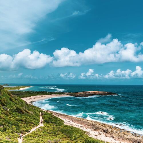 Mensen wandelen op een pad langs een begroeide rotsachtige kust voor een diepblauwe zee bij Pointe des Châteaux in Guadeloupe