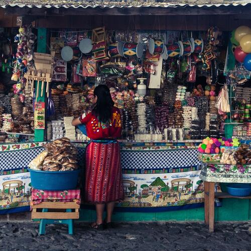 Een vrouw in felgekleurde traditionele kleding staat aan een rijkelijk gevuld marktkraam in Guatemala