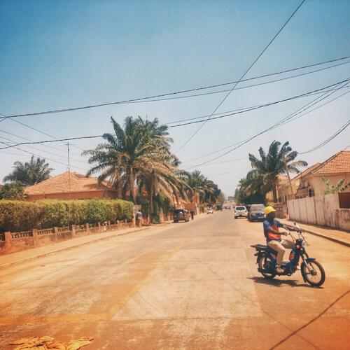Een man rijdt op een brommer tussen okergele huizen in Bissau