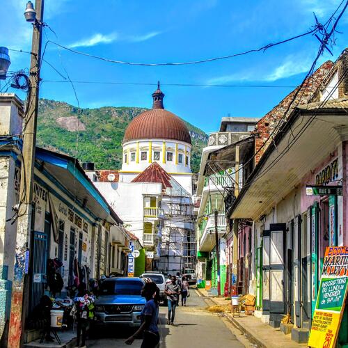 Mensen wandelen door een straat met felgekleurde koloniale gevels die leidt naar een grote kerk in Cap-Haïtien