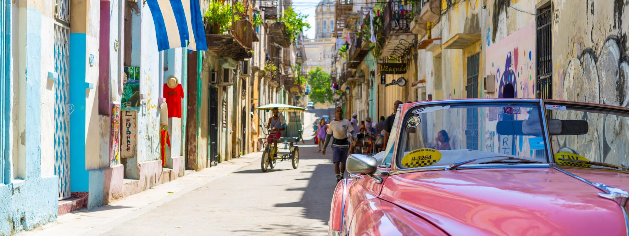 Een roze oldtimer staat in een smalle straat met oude huizen en een Cubaanse vlag in Havana, Cuba