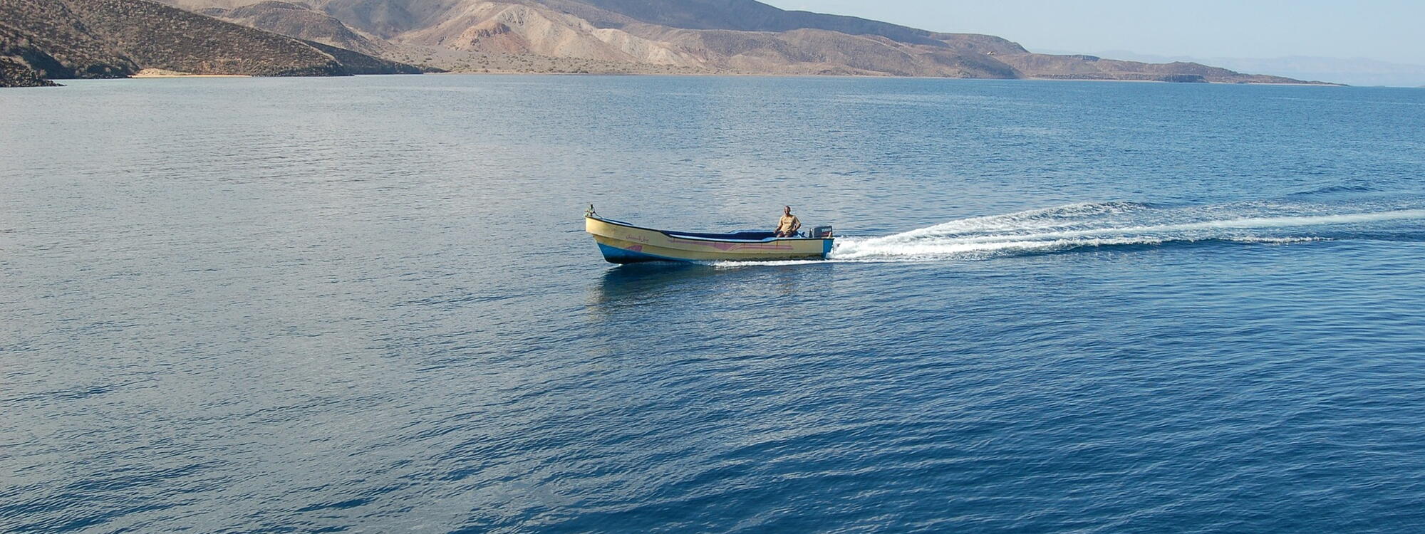 Een visser in een gele motorboot vaart voorbij de heuvelachtige kust van Djibouti