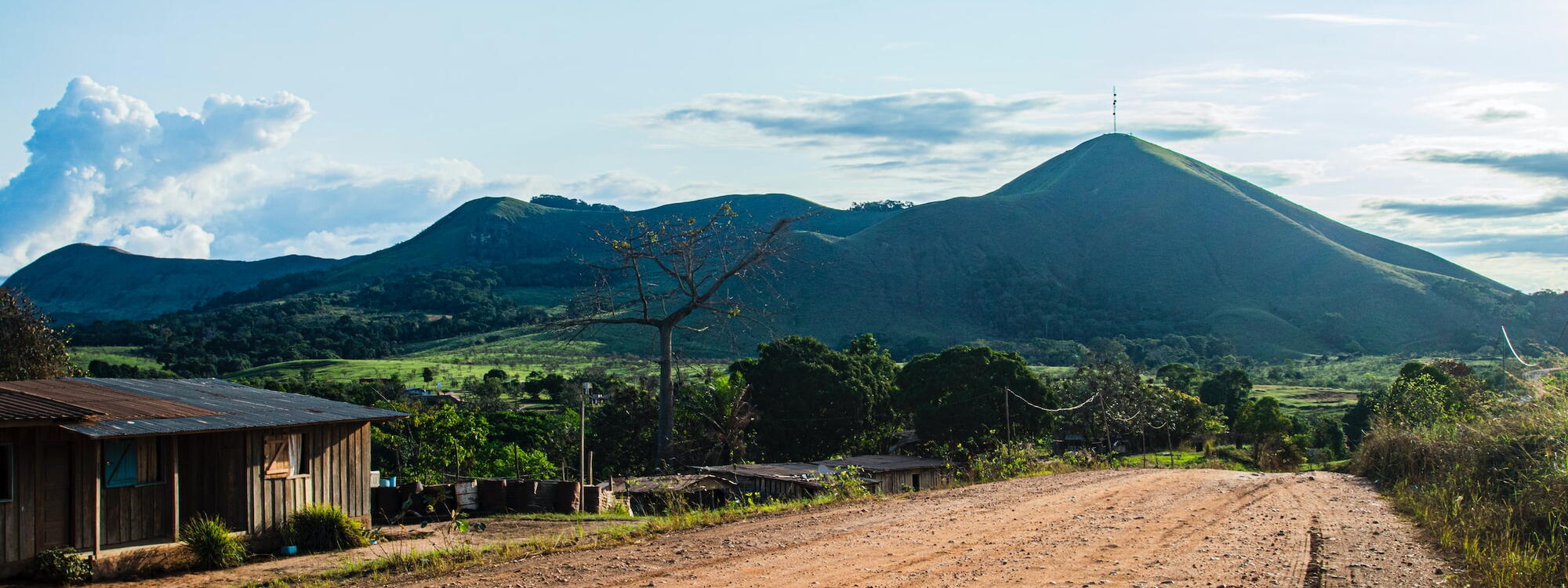 Een houten huisje staat langs een zandweg met op de achtergrond een grote heuvel nabij Lopé, in Gabon
