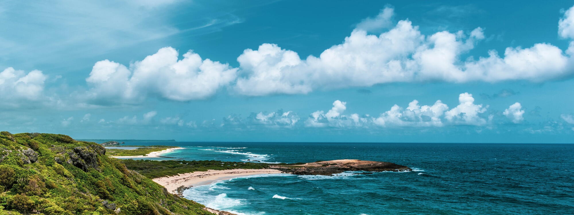 Mensen wandelen op een pad langs een begroeide rotsachtige kust voor een diepblauwe zee bij Pointe des Châteaux in Guadeloupe