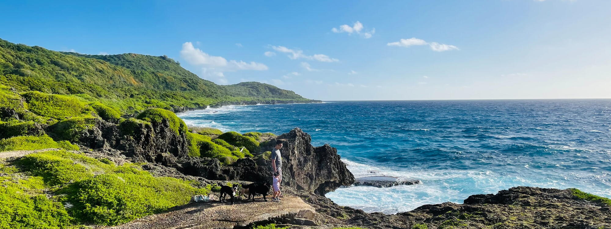 Een man wandelt met een kind en drie honden langs de rotsachtige kust bij Pågat op Guam