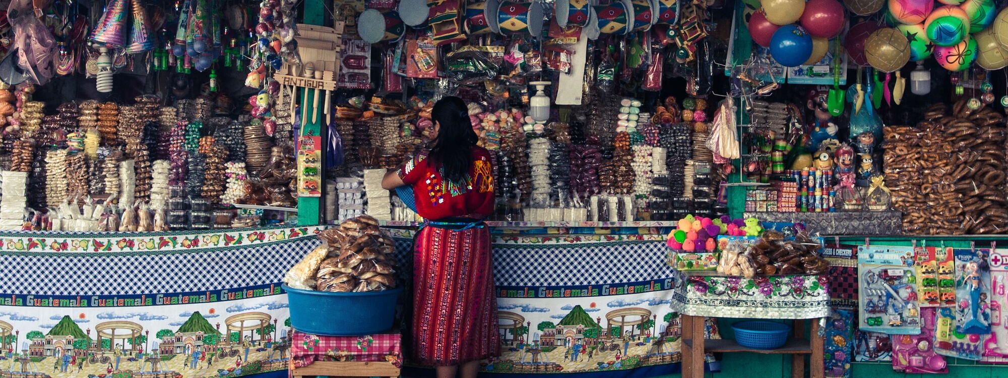 Een vrouw in felgekleurde traditionele kleding staat aan een rijkelijk gevuld marktkraam in Guatemala