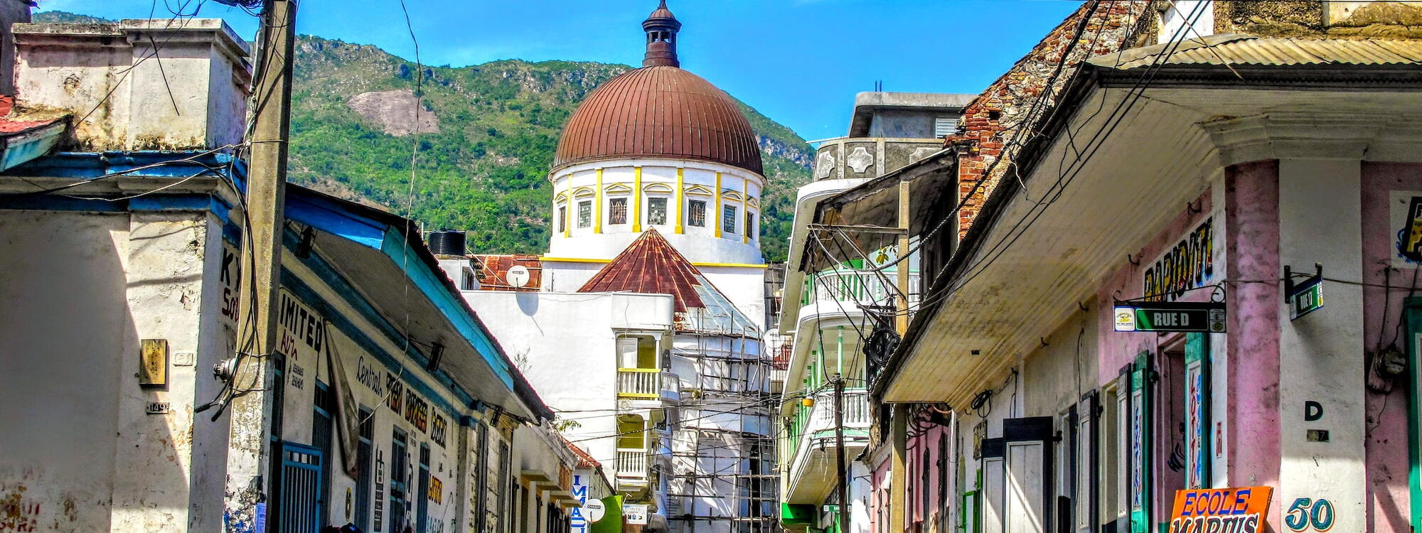 Mensen wandelen door een straat met felgekleurde koloniale gevels die leidt naar een grote kerk in Cap-Haïtien