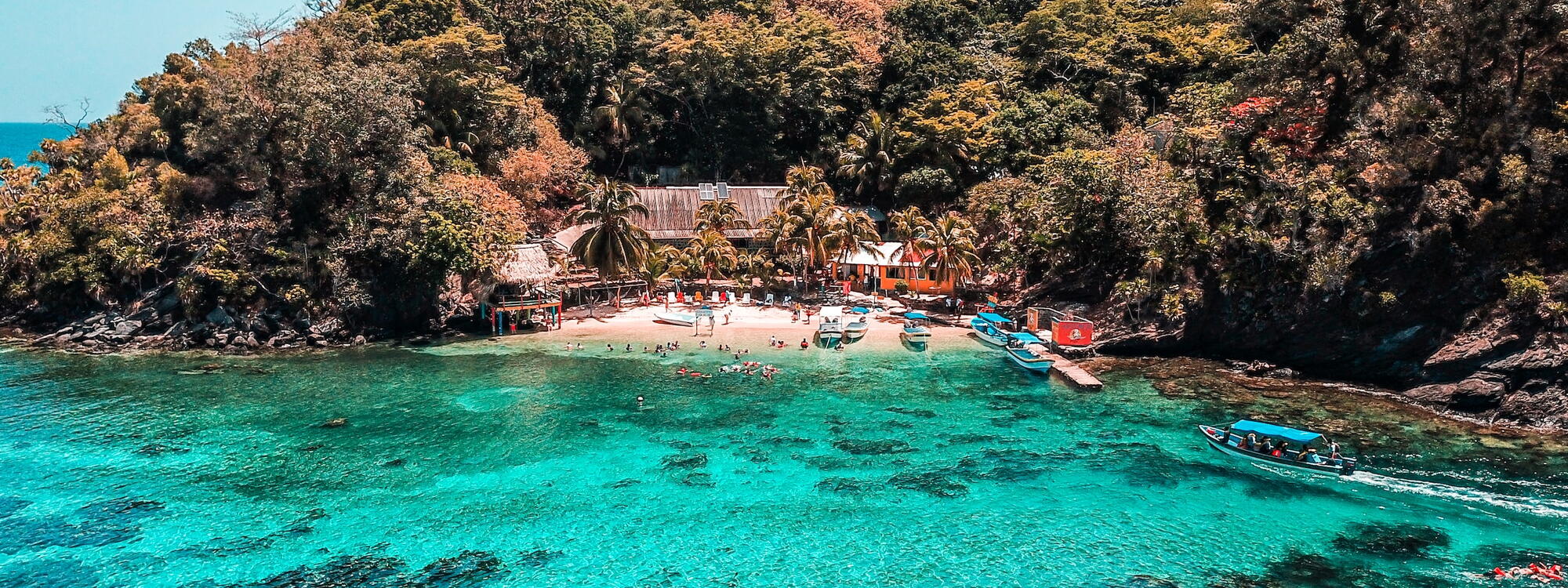 Een bootje vaart over het lichtblauwe water naar een klein strand dat verstopt ligt tussen de bomen en de rotsen op Cayos Cochinos in Honduras